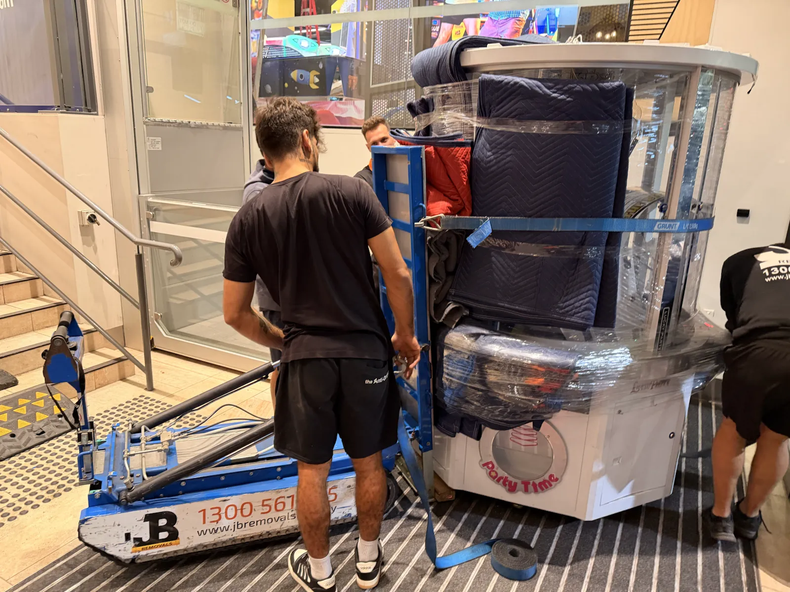 Three JB Removals crew members controlling a protected glass arcade machine on the Hercules stair climber