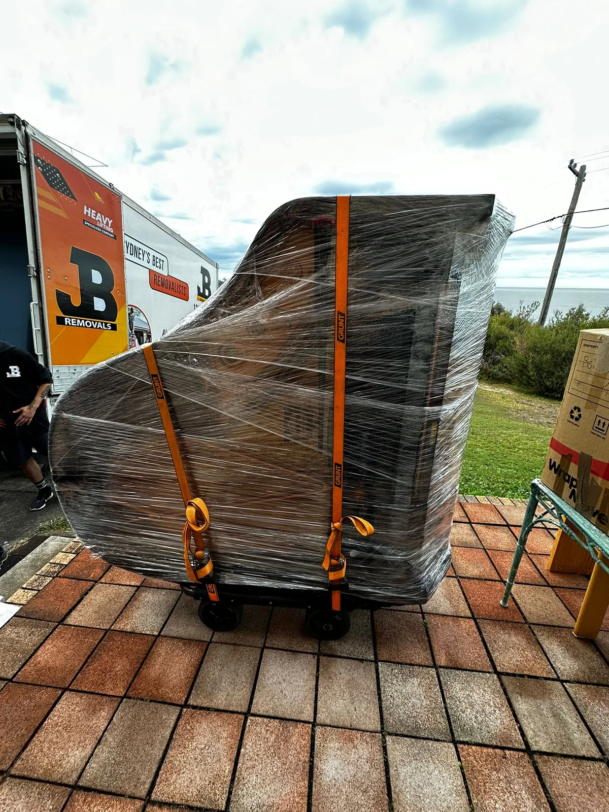 Wrapped upright piano secured on a trolley beside a JB Removals truck