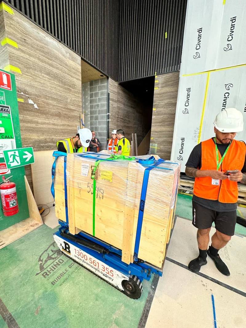 Timber crate secured with blue and green restraints on JB tracked handling equipment at a Sydney commercial site.