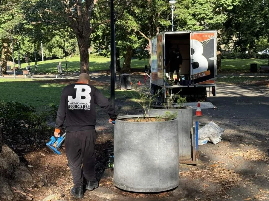 Large planted cylindrical pots beside a JB truck with a crew member carrying a blue hydraulic jack
