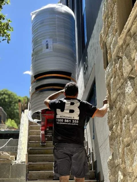 JB removalist guiding a wrapped heating vessel on a red stair-climbing trolley beside the building
