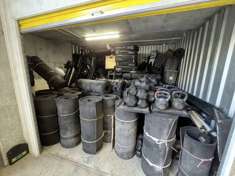 Stored gym equipment including rowers, dumbbells, kettlebells and rolls of flooring