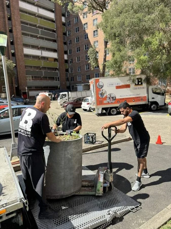 Three JB crew members handling a large cylindrical planted pot on a truck tail lift with a pallet jack