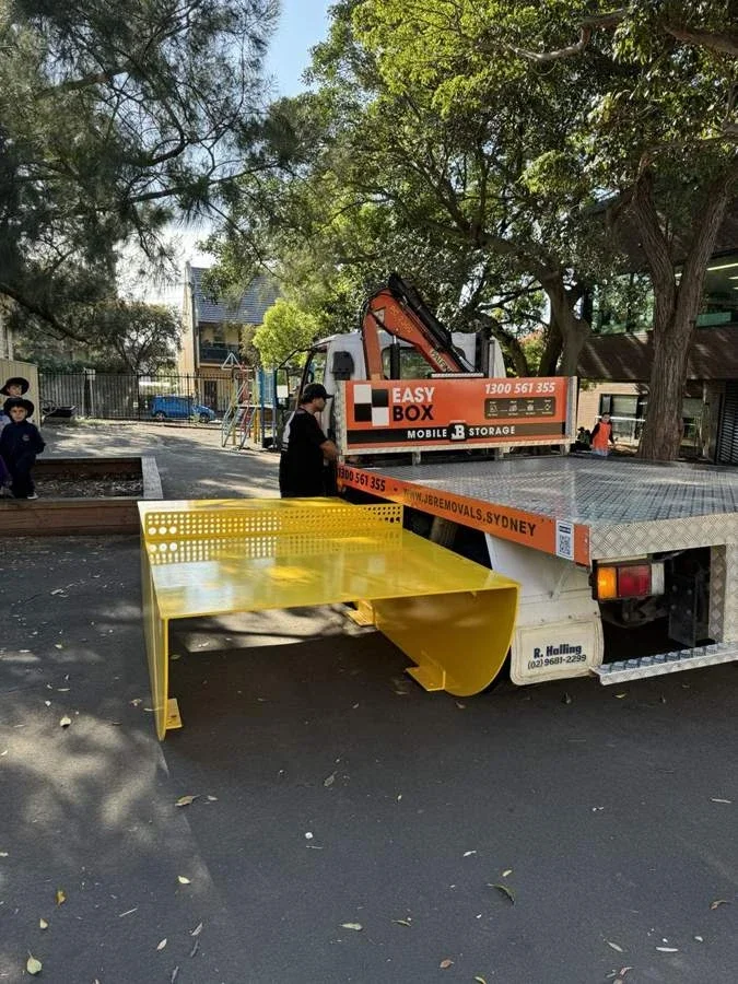 Yellow metal table-tennis table beside the rear of a JB EasyBox truck at an outdoor recreation area