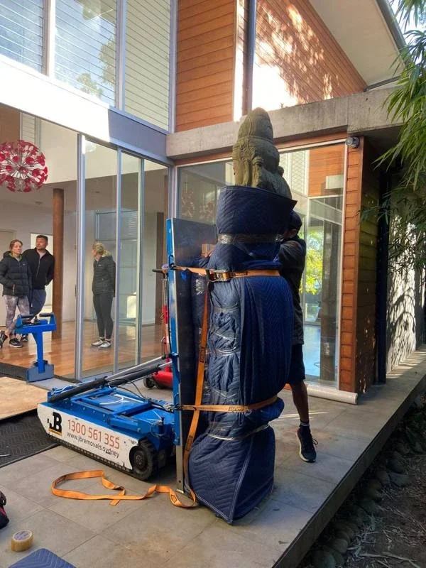 Tall garden statue padded with blue removal blankets and secured with orange straps beside powered moving equipment