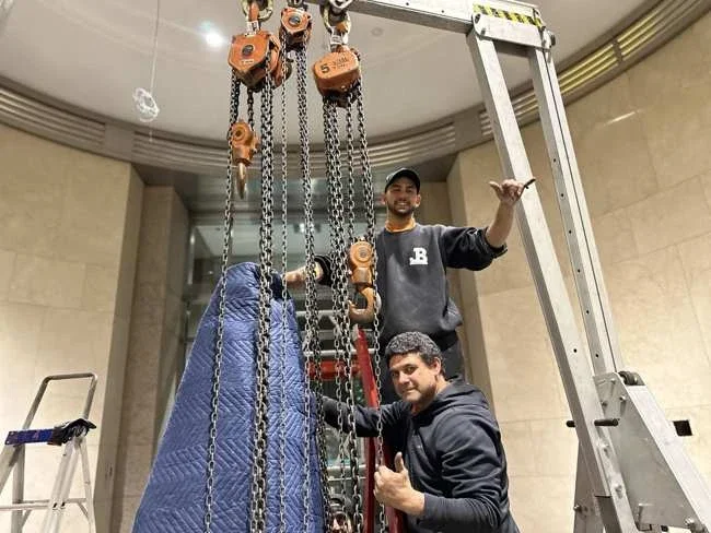 JB team beside a padded sculpture section beneath a gantry and chain hoists