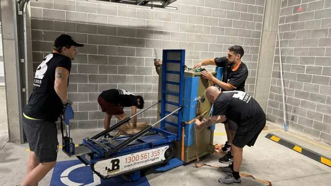 Team beside a large strapped safe on blue handling equipment in a garage