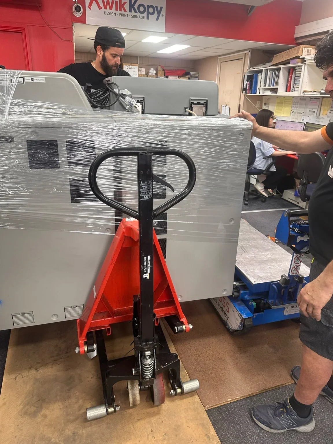 JB crew beside a wrapped printer, pallet jack and blue stair climber during loading