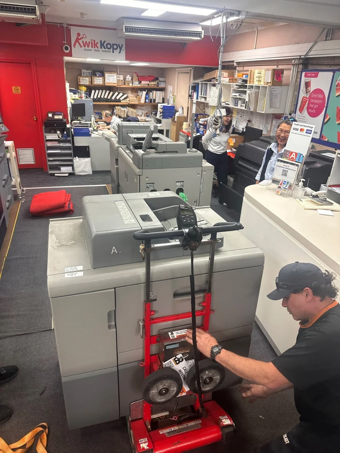JB team member adjusting a red stair climber beside a strapped printer at the print premises