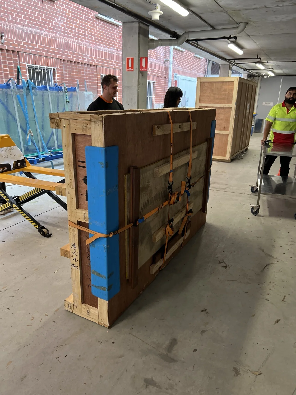 Timber transport crate secured with orange straps and blue protective padding in a covered campus access area