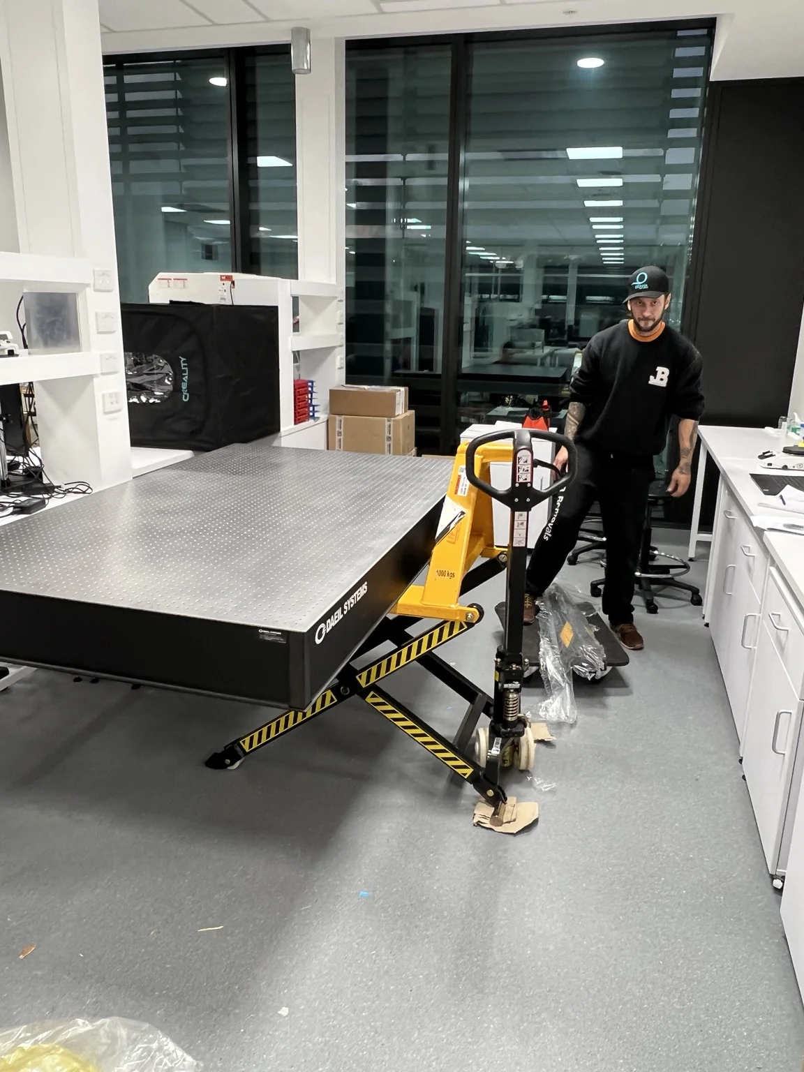 DAEIL SYSTEMS optical table raised on a high-lift scissor pallet jack beside a JB removalist