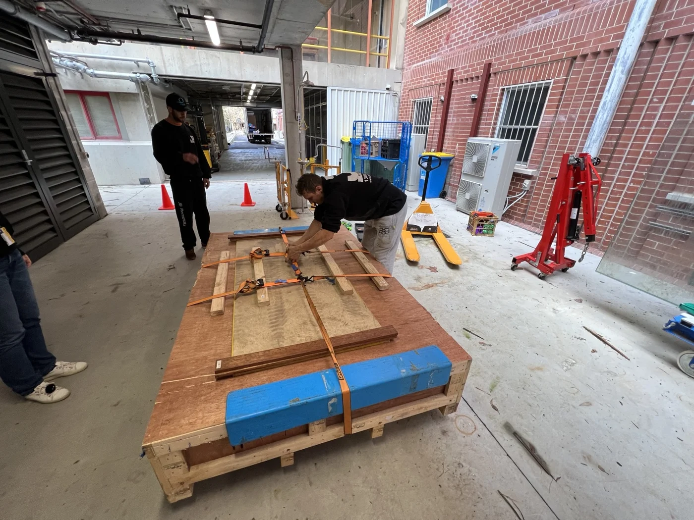 JB crew preparing a strapped timber crate beside a pallet jack and portable hydraulic workshop crane