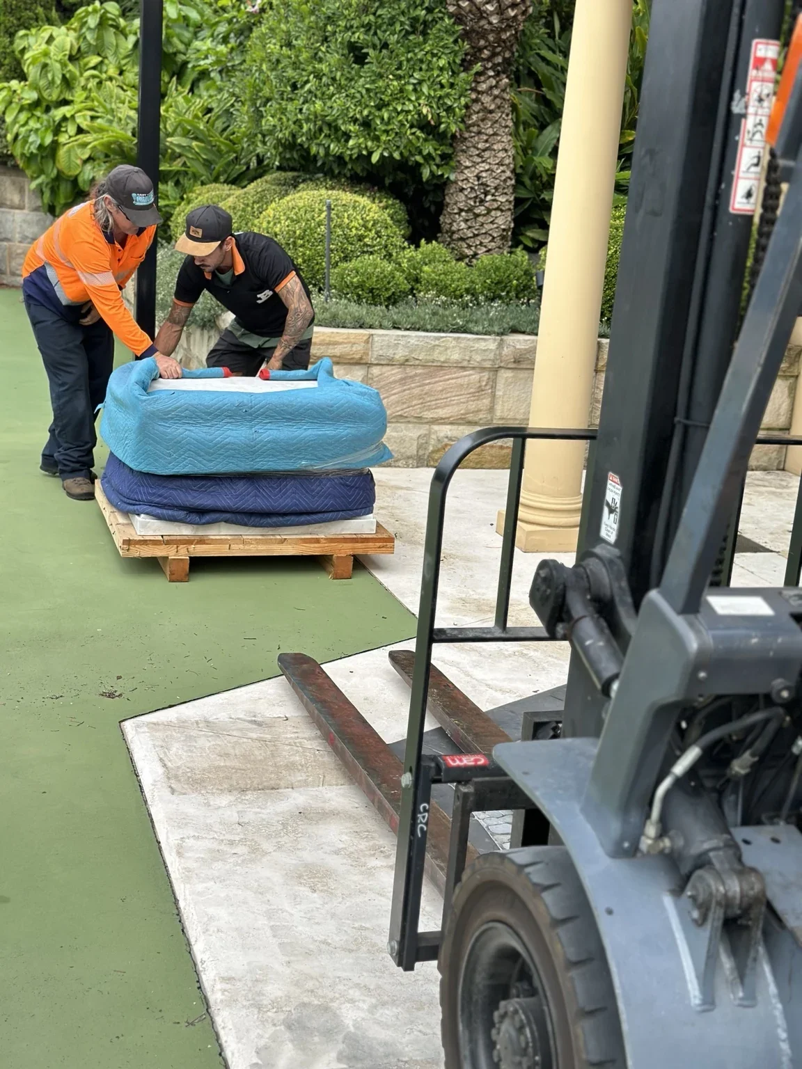 JB crew beside a padded marble base on a pallet with a forklift nearby