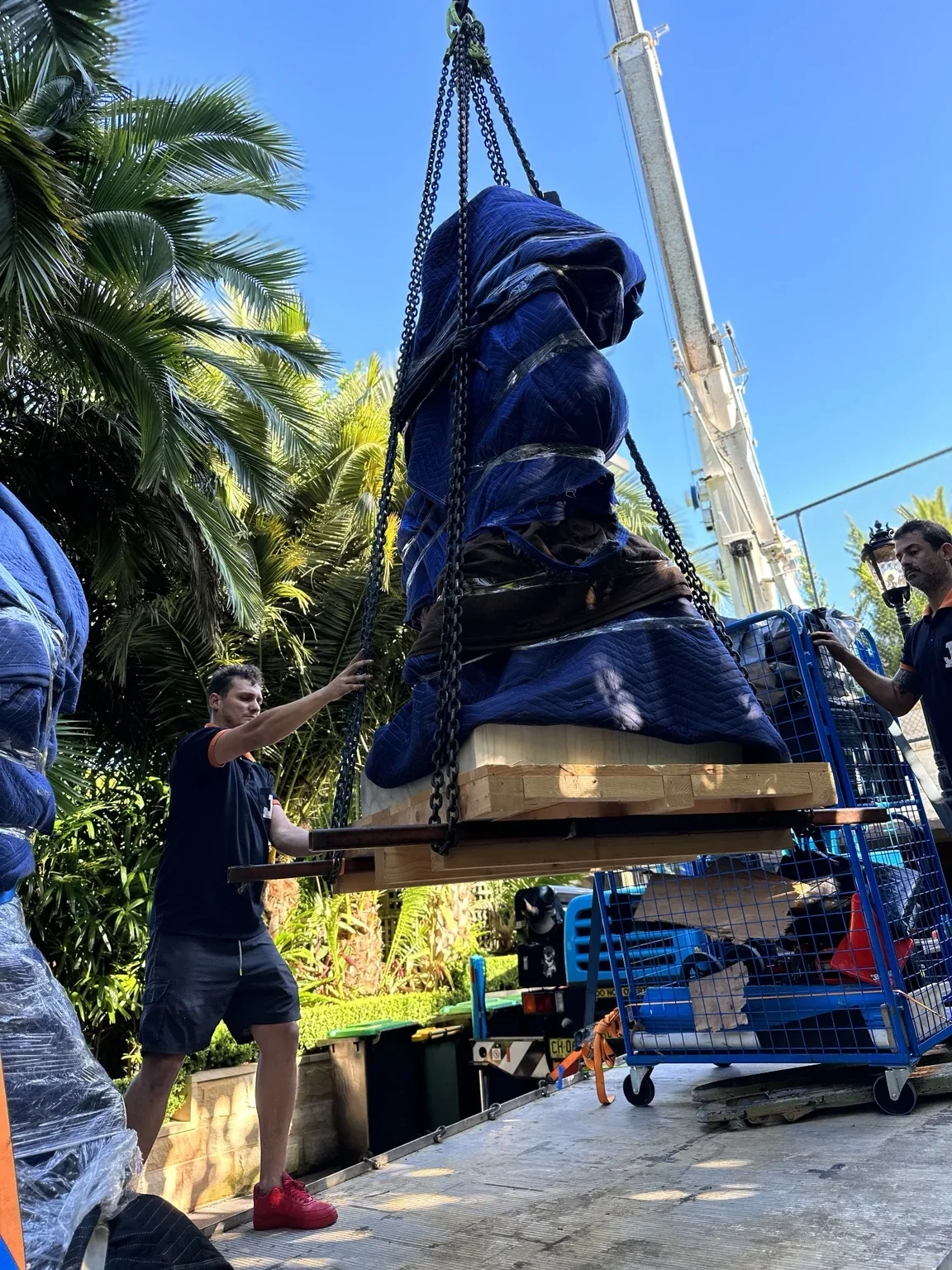 JB crew beside a blanket-wrapped statue suspended on a pallet by a crane