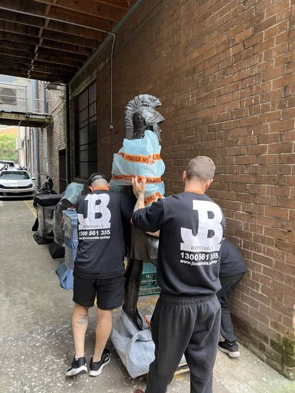 JB removalists guiding a padded bronze warrior sculpture along a brick-walled external passage