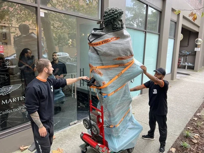 JB removalists with a padded and strapped bronze sculpture on red moving equipment outside the collection showroom