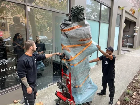 JB Removals: JB removalists with a padded and strapped bronze sculpture on red moving equipment outside the collection showroom